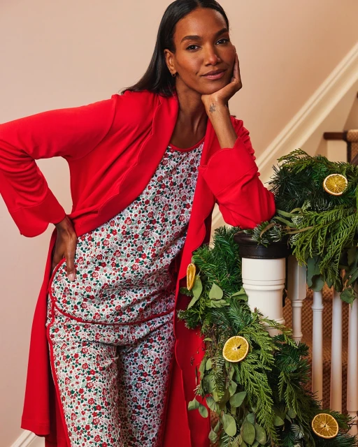 Elegant dark-haired woman in red robe and floral pajamas leaning on staircase banister with festive evergreen garland and orange slices.