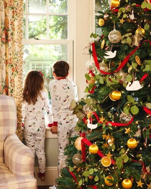 A young girl and boy in pajamas look out a window next to a Christmas tree decorated with gold ornaments and red ribbon in a home interior.