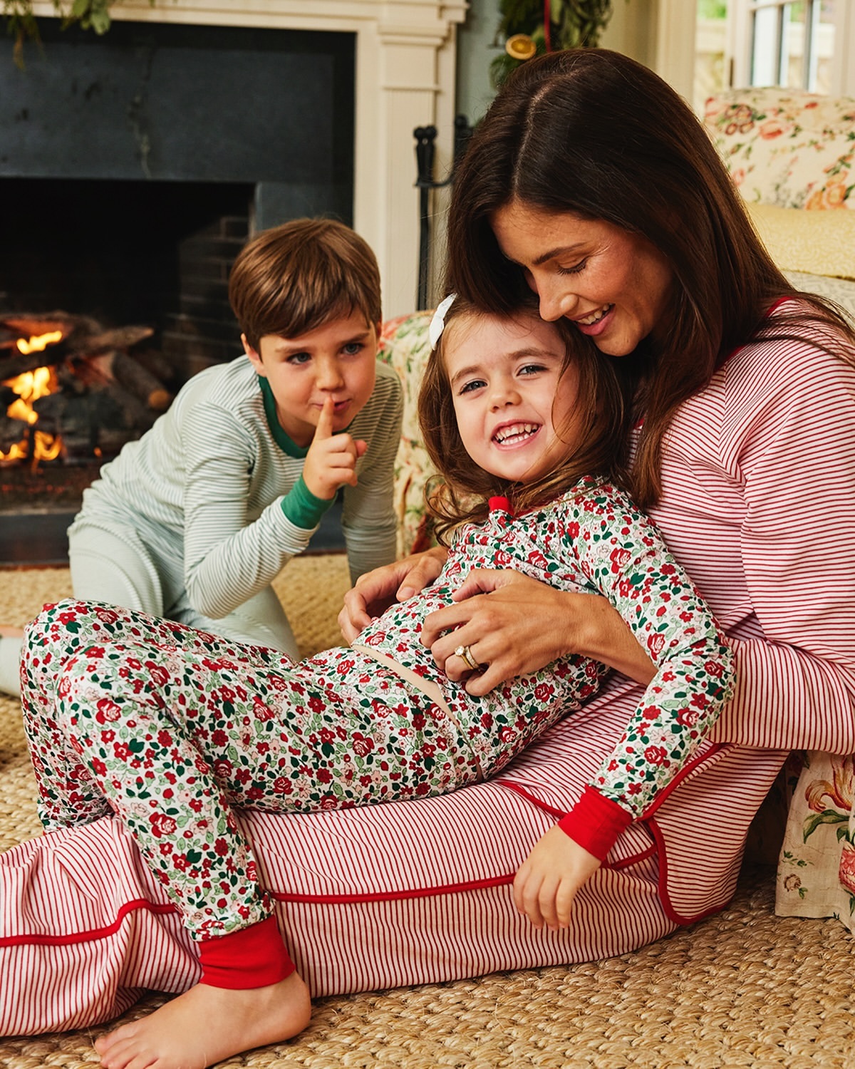 A smiling mother is sitting on the floor with her daughter in pajamas, as her son stands in the background near a fireplace.