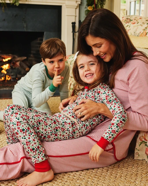 A smiling mother is sitting on the floor with her daughter in pajamas, as her son stands in the background near a fireplace.
