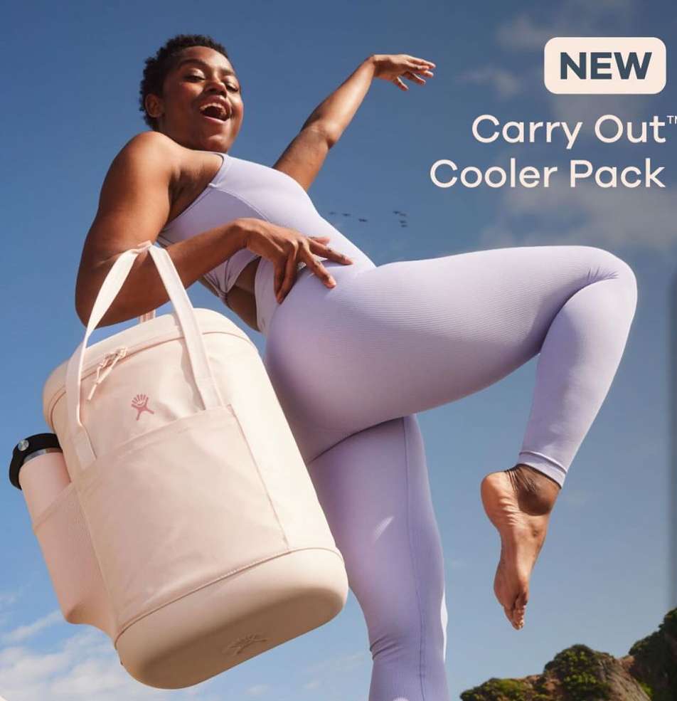 A woman in activewear poses outdoors with a Hydro Flask cooler pack, smiling against a blue sky, showcasing fitness and travel lifestyle.