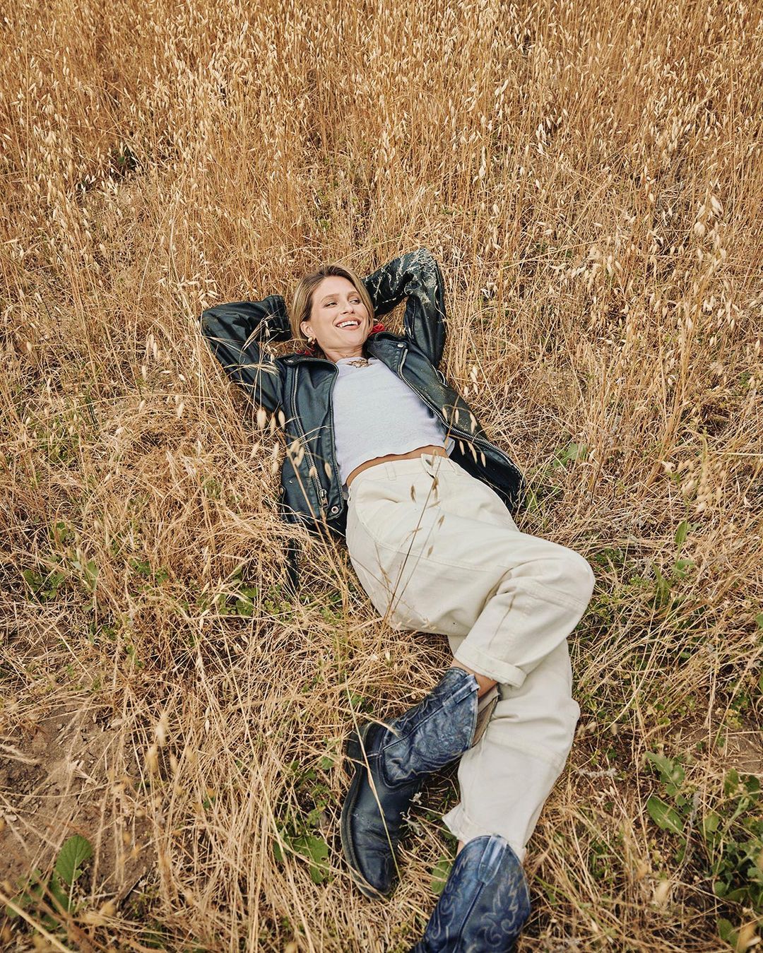 Young woman lying in a field of tall, dry grass, wearing a black leather jacket, white top, light pants, and blue cowboy boots.