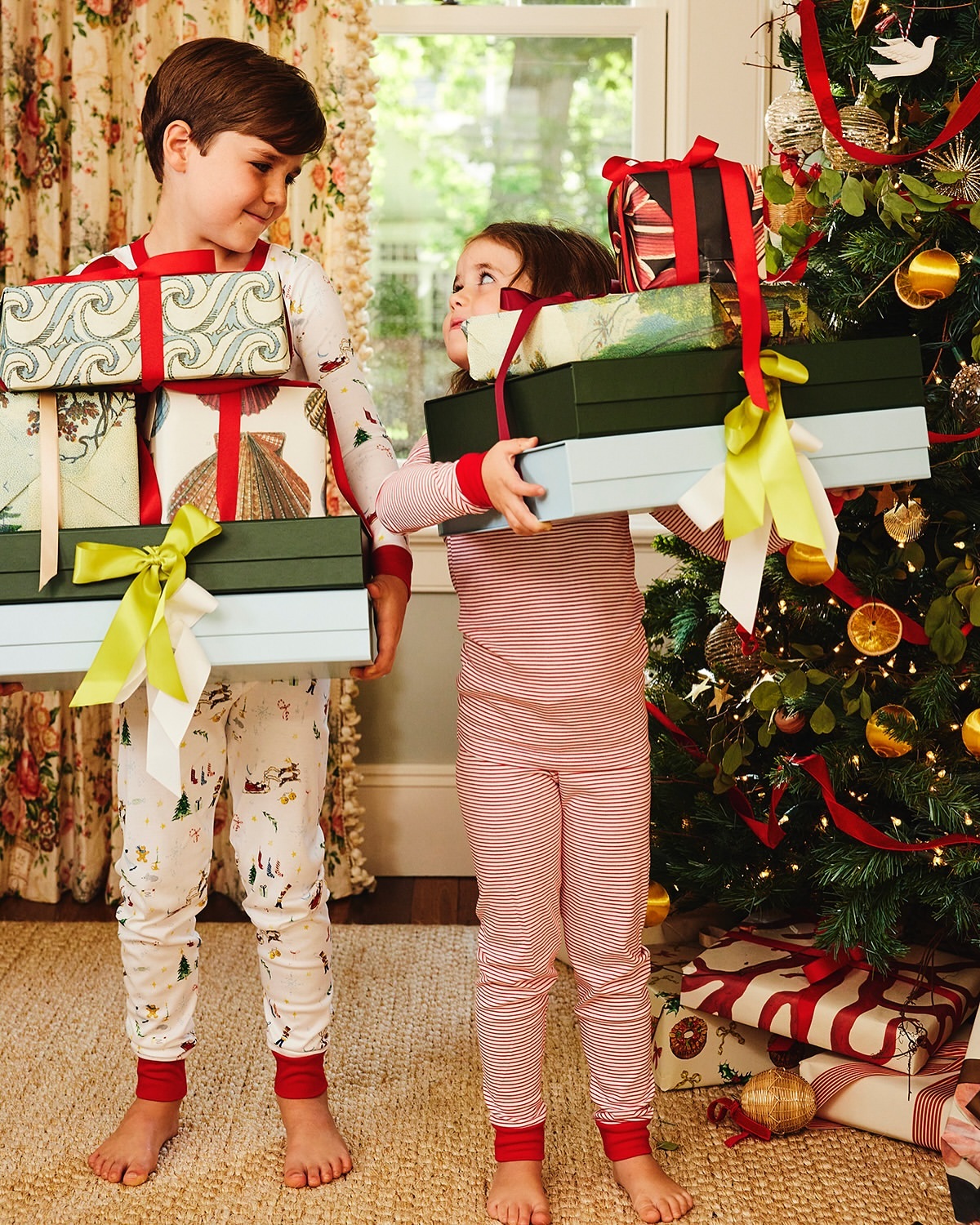 Two children in pajamas exchanging wrapped Christmas presents, standing near a decorated tree in a brightly lit indoor setting.