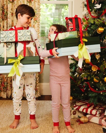 Two children in pajamas exchanging wrapped Christmas presents, standing near a decorated tree in a brightly lit indoor setting.