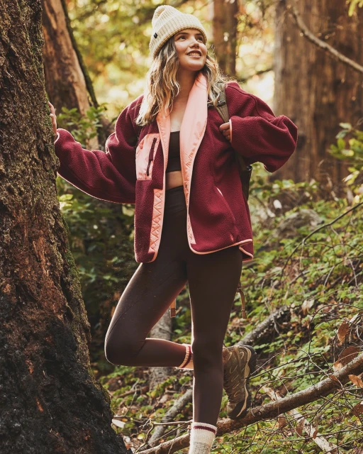 A woman in a red fleece jacket and brown leggings poses near a tree in a forest setting, looking up and to the side, styled for outdoor lifestyle.