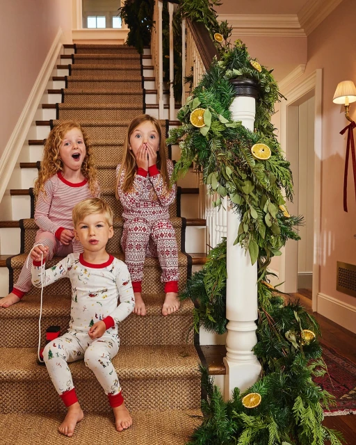 Three children in pajamas sitting on a staircase decorated with garland, creating a festive and joyful holiday atmosphere in an indoor setting.