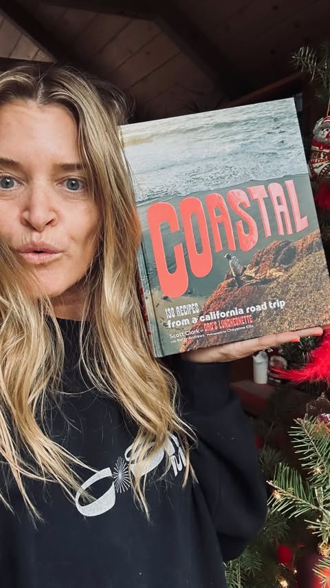 Woman holding a cookbook titled 'Coastal' in front of a decorated Christmas tree. Cookbook features a coastal scene and red text.