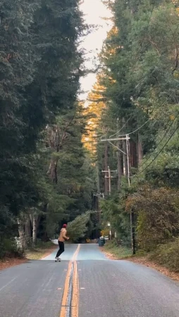 A person longboarding down an asphalt road lined with tall trees in a forest setting, enjoying an outdoor recreation activity.