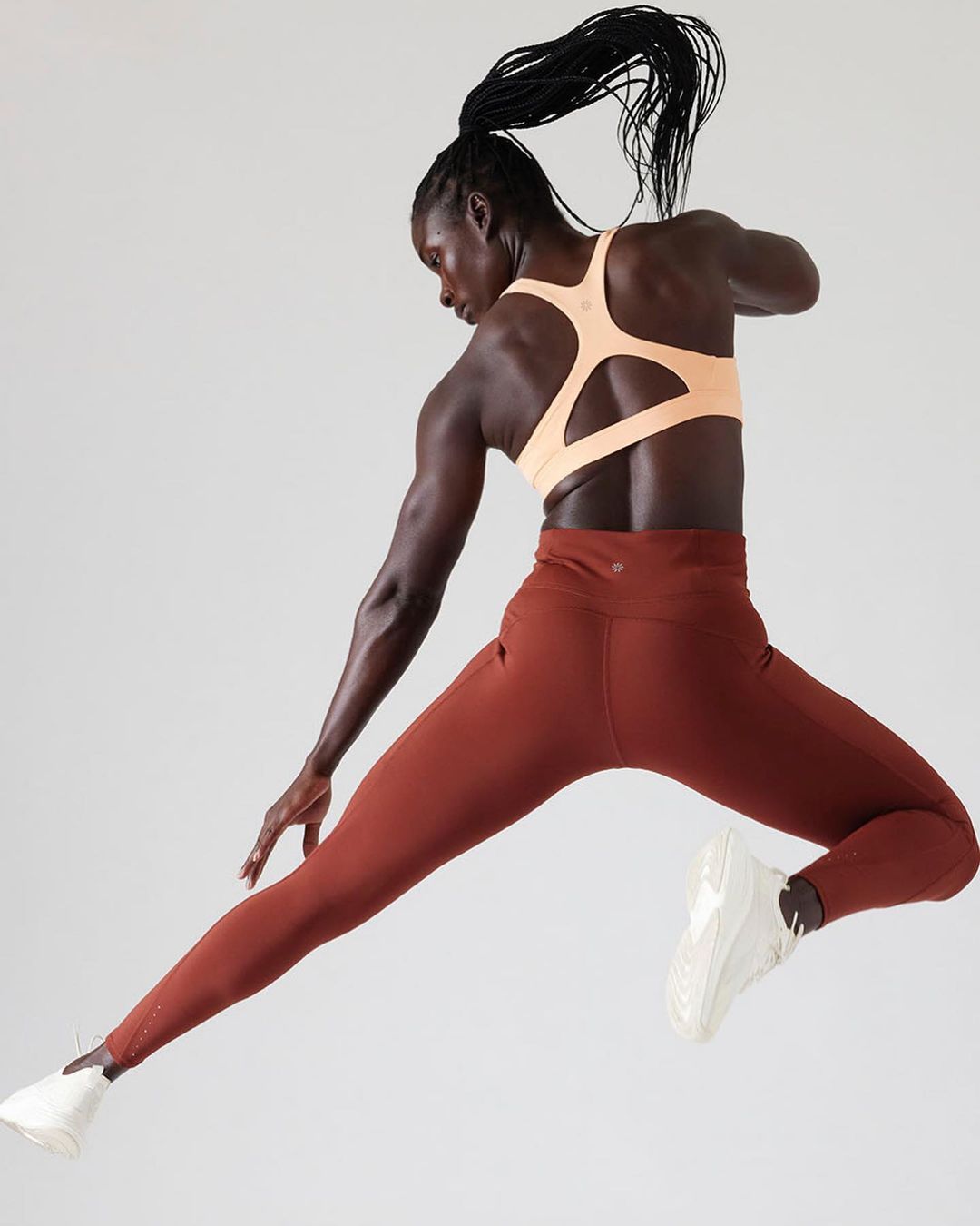 Black woman in a peach sports bra and rust leggings captured mid-leap against a clean white backdrop by Cheyenne Ellis.