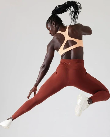Black woman in a peach sports bra and rust leggings captured mid-leap against a clean white backdrop by Cheyenne Ellis.