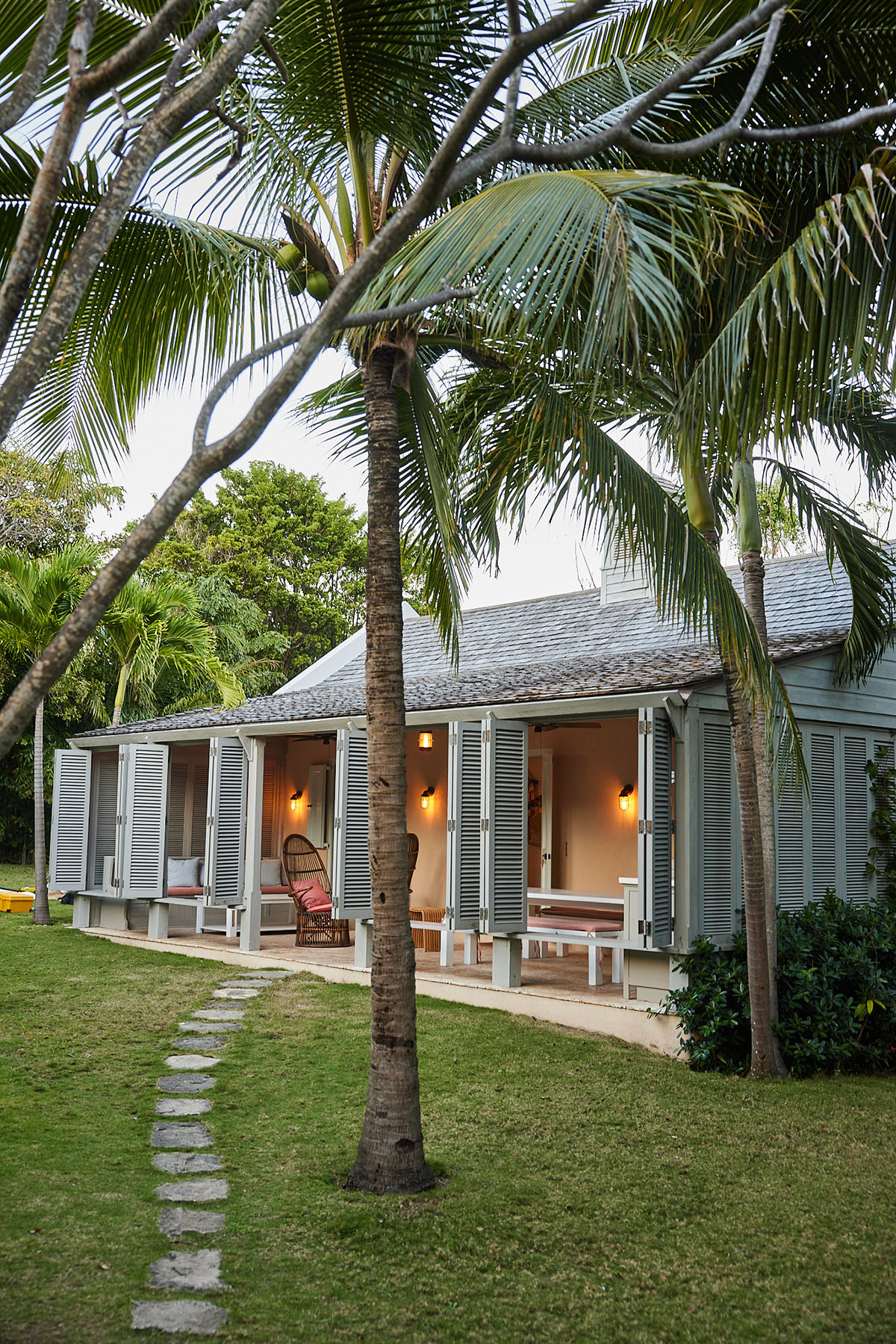 Exterior view of a gray wooden house with palm trees, a stone pathway, and tropical foliage on a sunny day in a tropical location.
