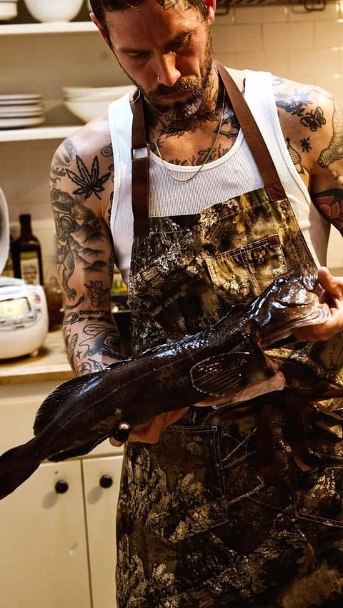 Adult male in apron holding fish indoors. The man has tattoos. Kitchen shelves are in the background.
