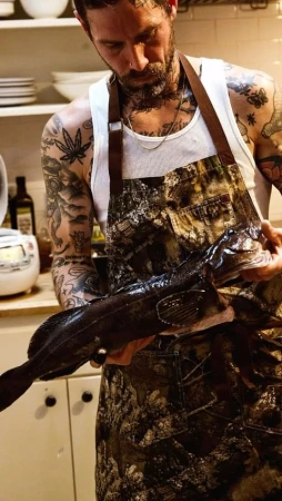 Adult male in apron holding fish indoors. The man has tattoos. Kitchen shelves are in the background.
