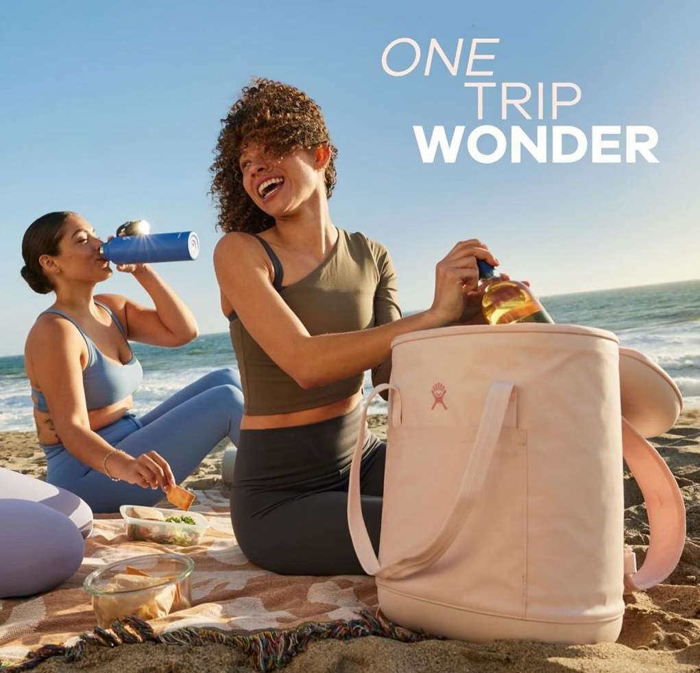 Two women on a beach, one opening a Hydro Flask cooler, the other drinking from a bottle, with food containers visible on a patterned blanket.