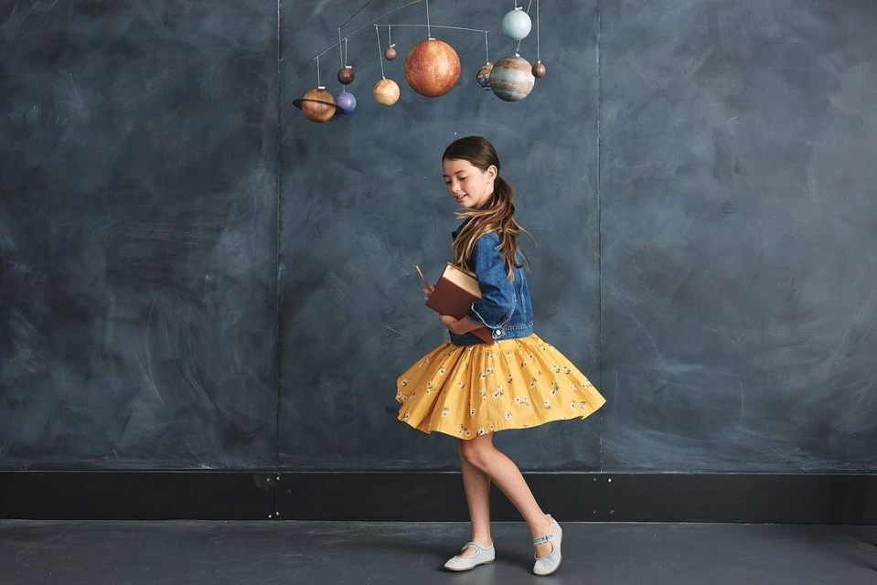 Young girl in a yellow skirt holds a book, walking in front of a gray wall with hanging planet models for a children's fashion shoot.