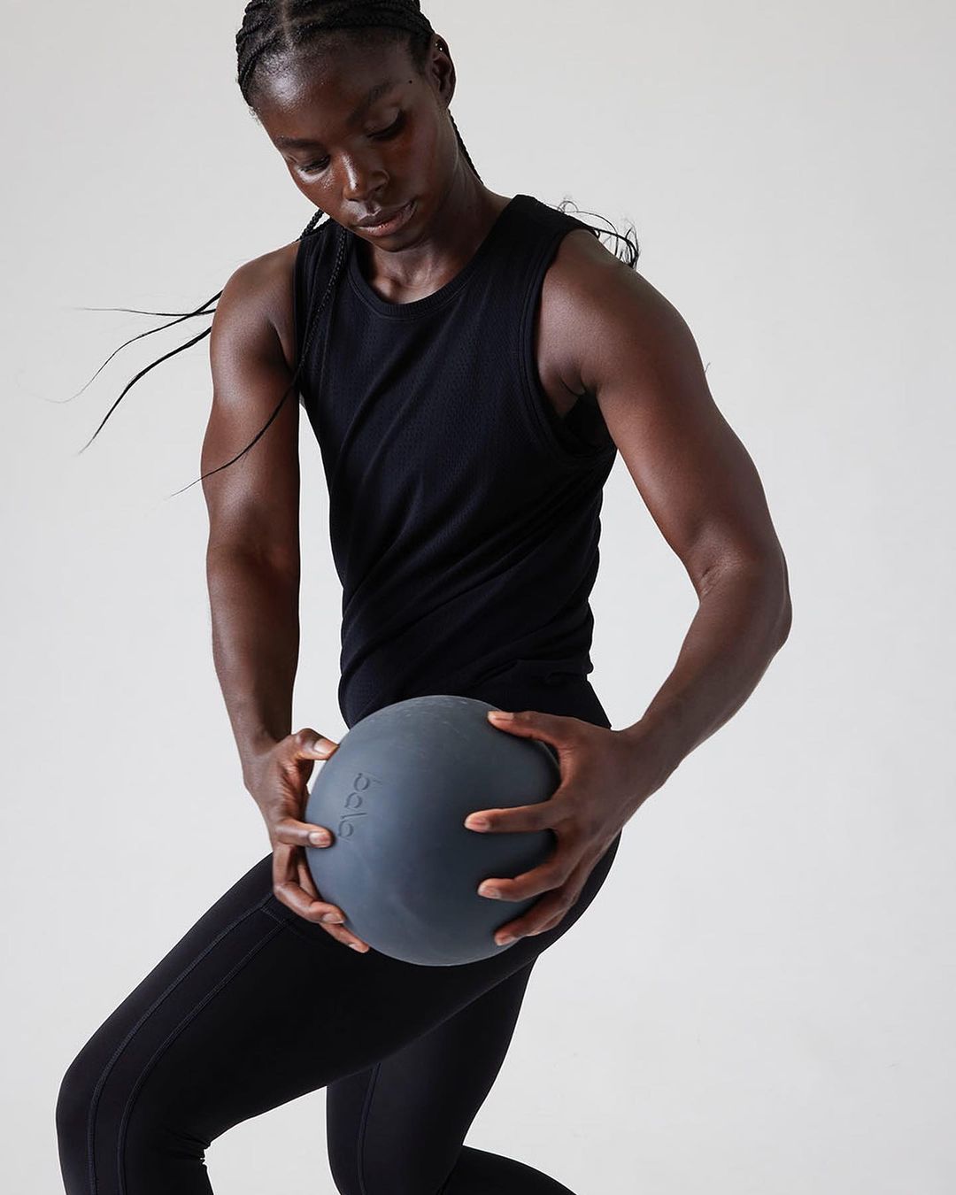 Athletic African American woman in fitness apparel holding a grey exercise ball in a bright indoor studio setting, promoting fitness.