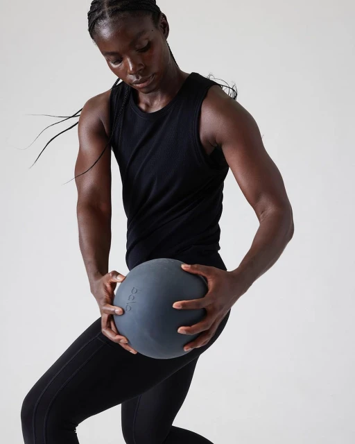 Athletic African American woman in fitness apparel holding a grey exercise ball in a bright indoor studio setting, promoting fitness.