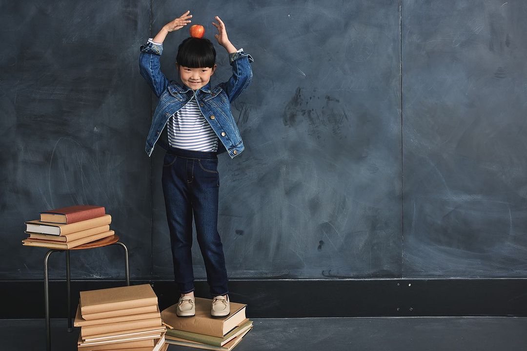 A young girl balancing an apple on her head smiles while standing on a stack of books in front of a dark blue textured background.