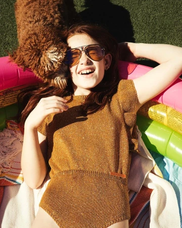 Young girl wearing aviator sunglasses and gold knit romper lying in a colorful inflatable pool with a brown dog