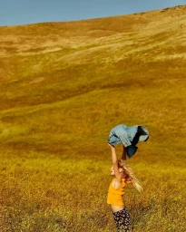 Young girl in yellow top holding up a denim jacket in a grassy meadow under warm sunlight