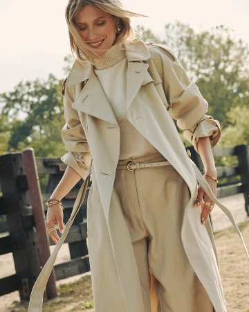 Woman in beige trench coat and matching trousers smiling outdoors near a dark wooden fence
