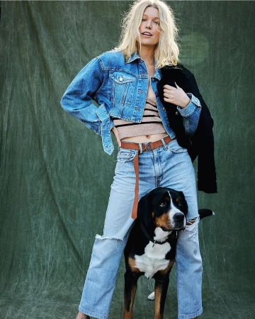 Blonde woman wearing denim jacket and jeans standing with a large dog against green textured backdrop