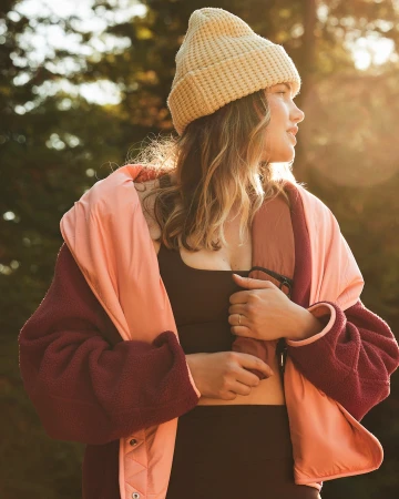 Woman wearing a cream knit beanie and a burgundy fleece jacket outdoors during golden hour sunlight.