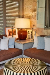 Interior corner setup featuring brown low-profile sofas with blue and white striped cushions, a large amber glass lamp, and a patterned round coffee table.