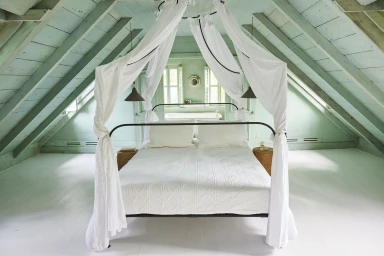 A canopy bed with white draping fabrics under a pale green wooden slanted attic ceiling with exposed beams.