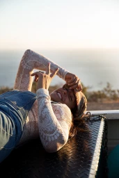 Smiling woman in a sheer lace top and denim overalls lying on the back of a truck during sunset.