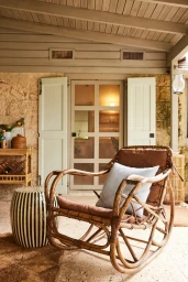 A rattan rocking chair with brown cushions and a light blue pillow on a covered outdoor porch with stone walls.