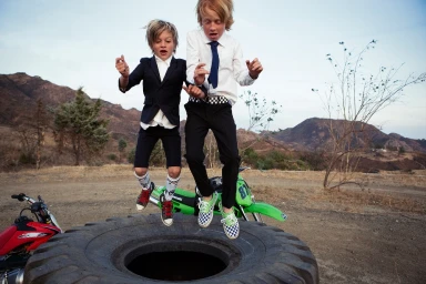 Two boys in suits jumping in the air in front of a dirt landscape with a green dirt bike and a large tire