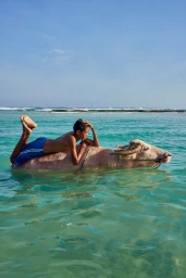 Young boy lying prone on the back of a light-colored water buffalo wading in clear blue coastal water with breaking waves.
