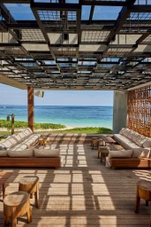 Open-air patio with wooden decking, plush seating, and a patterned overhead trellis casting geometric shadows facing the ocean.