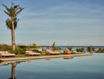 Resort pool terrace featuring lounge beds on green grass, palm trees, and ocean views under a clear sky.