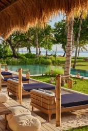 Outdoor resort lounge area with rattan daybeds and dark blue cushions facing a swimming pool and ocean view under a thatched roof.