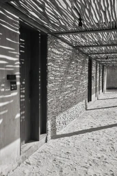 Monochrome architectural corridor with stacked stone walls, sandy ground, and an intricate slatted wooden overhead canopy casting patterns.