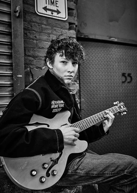 Black and white portrait of a young adult male musician with curly hair playing an electric guitar in an urban setting.