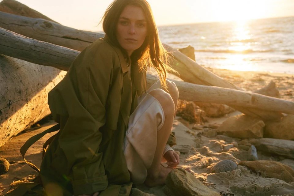 A woman in a long olive coat poses on a beach with driftwood and rocks at golden hour, looking directly at the camera in a natural setting.