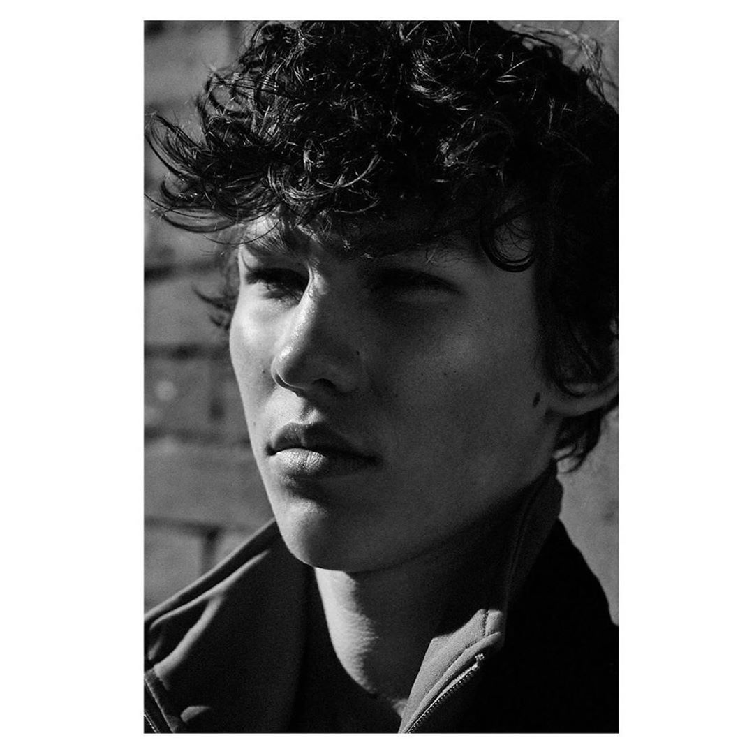 Close-up monochrome portrait of a young man with dark curly hair and a serious expression by photographer Squire Fox.