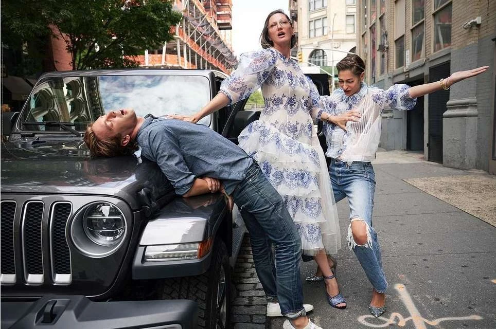 Three people pose with a Jeep on a city street. A blond man lies on the hood. Two women in dresses pose beside him.