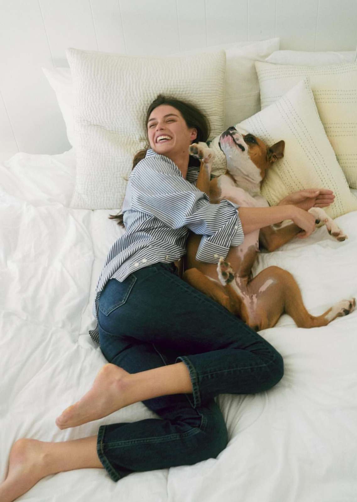 Smiling woman in jeans and a striped shirt lies on a bed with pillows, hugging her brown and white dog.