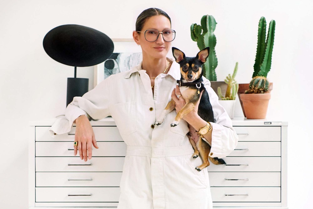 Adult woman poses with small dog in white jumpsuit. Behind them is a white cabinet, sculpture, houseplants, and neutral decor.
