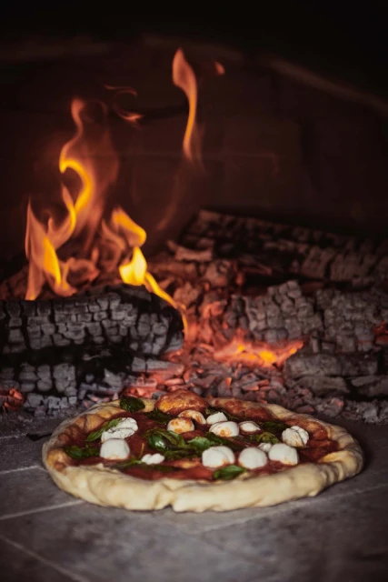Overhead shot of a wood-fired pizza with mozzarella and basil, presented inside the brick oven with flames and charred wood.