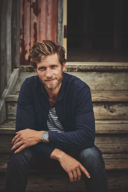 Blond male adult sits on wooden steps in outdoor shot with a casual navy sweater, striped shirt, watch, and rustic door in the background.