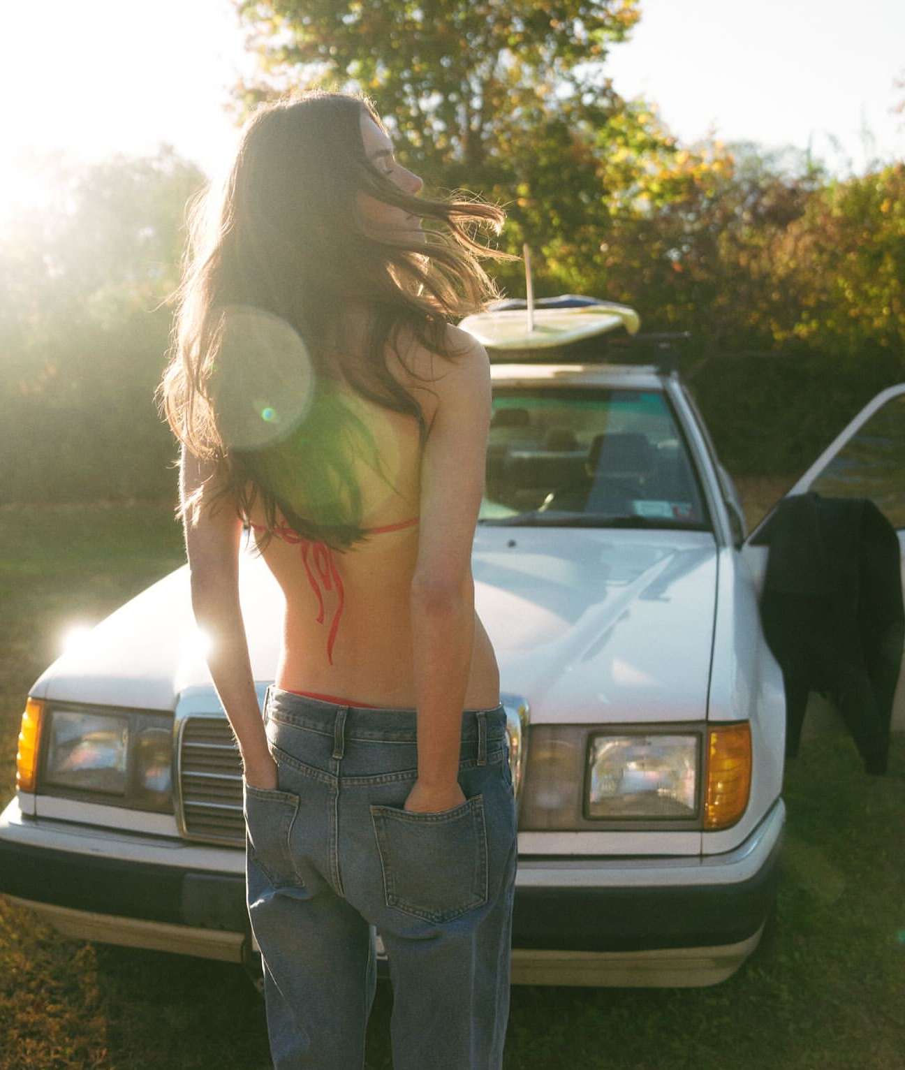 Woman in bikini top and jeans stands by a white car with a surfboard on top against trees with sunlight flare, outdoor setting.