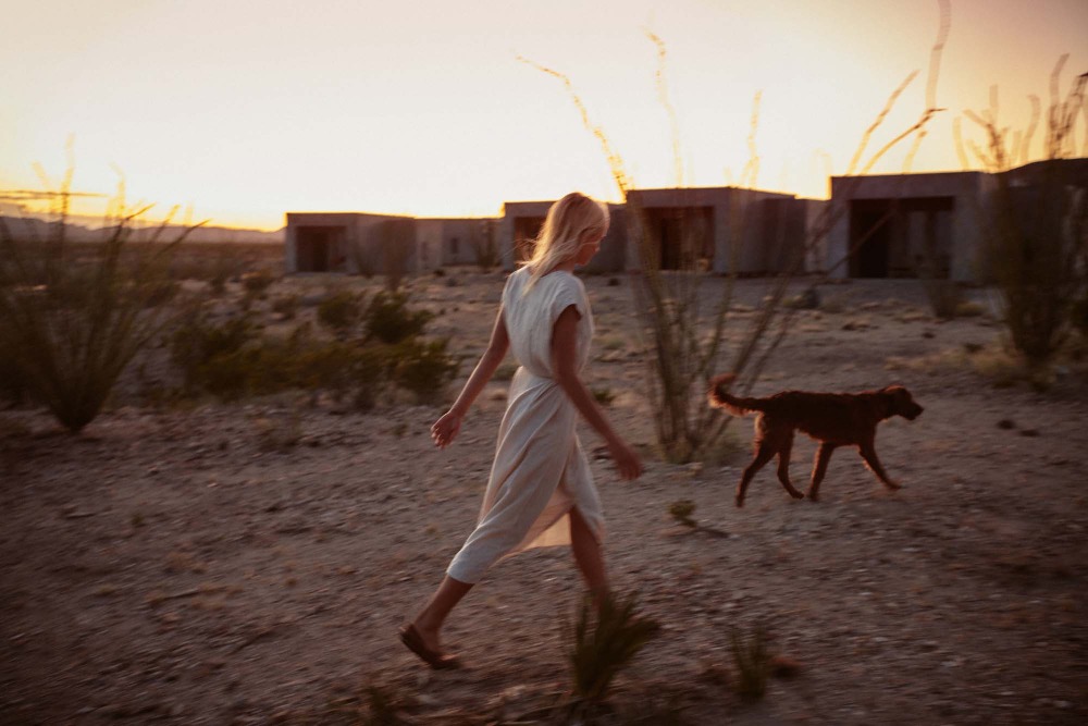 A woman in a dress walks a dog in a desert landscape at sunset with modern architecture in the background, creating a tranquil scene.