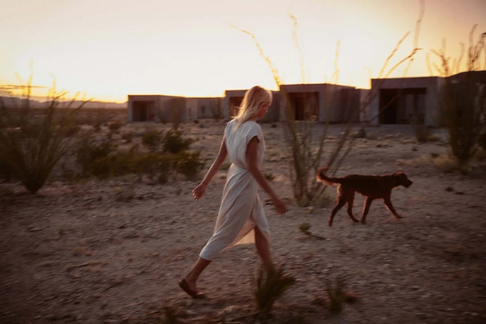A woman in a dress walks a dog in a desert landscape at sunset with modern architecture in the background, creating a tranquil scene.