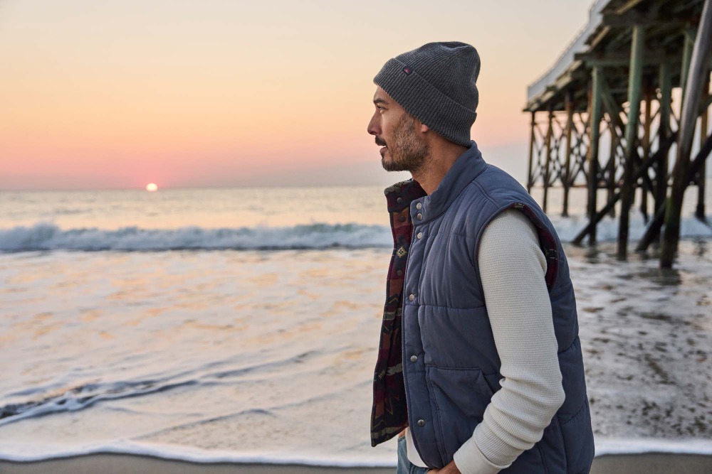 Man in vest and beanie on beach looks at sunrise over ocean with pier supports in background. Casual style for lifestyle content.
