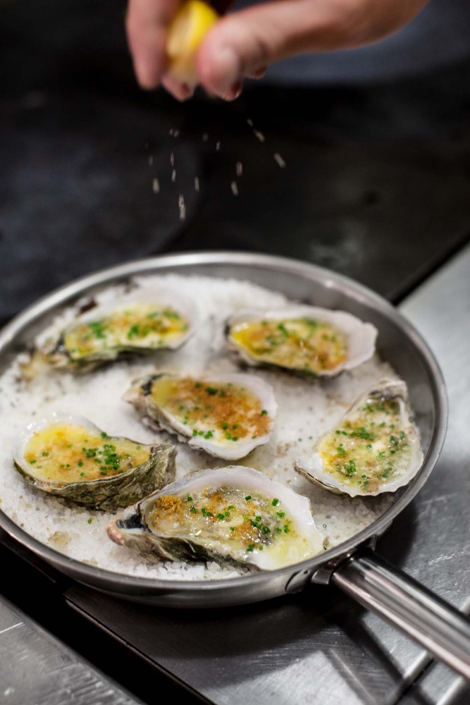 Overhead shot of a hand squeezing a lemon over baked oysters served on a bed of salt in a silver pan in a restaurant kitchen.